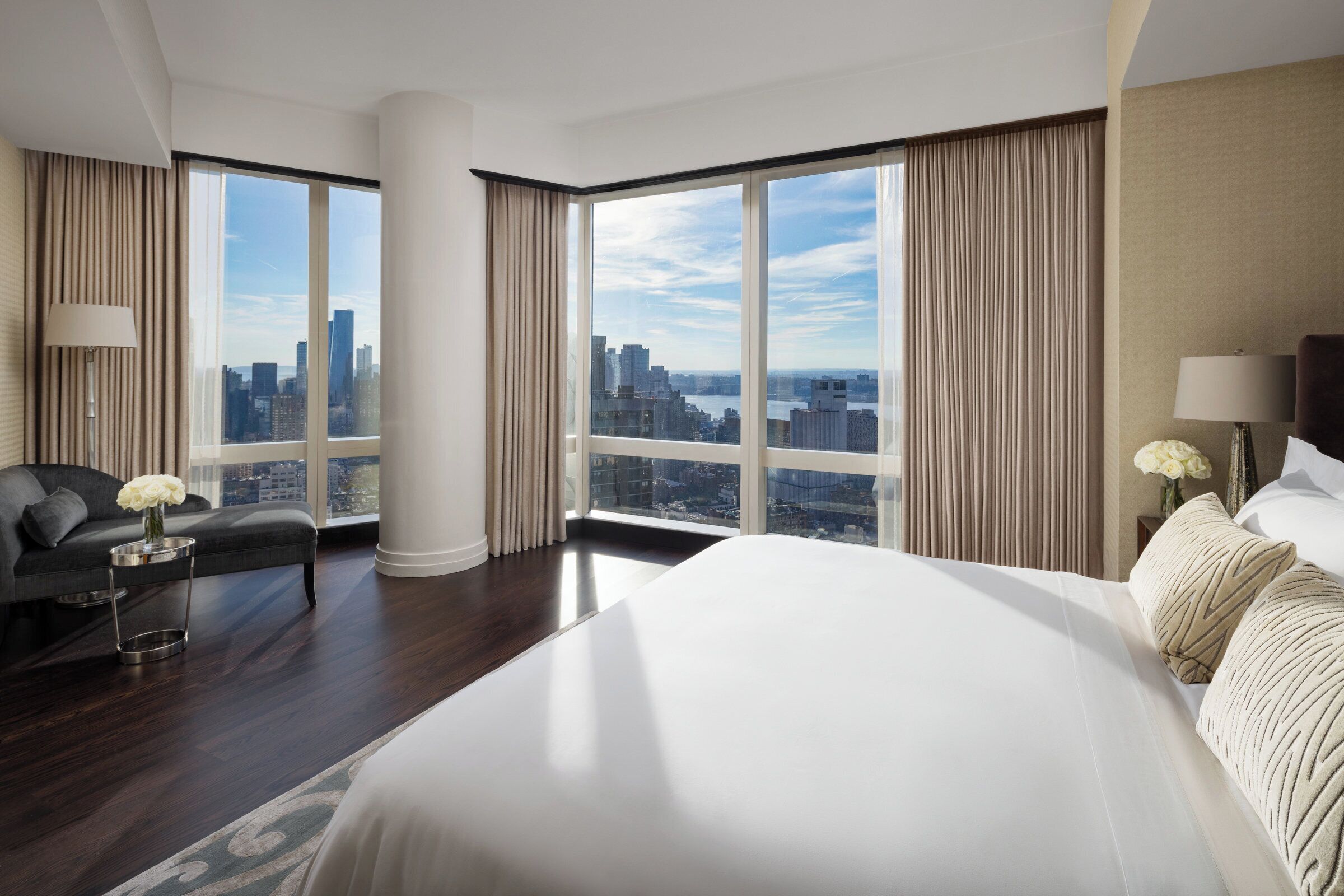 Een moderne hotelkamer met een groot bed, een uitzicht op de skyline doorgaande raampartijen en een donkere houten vloer.