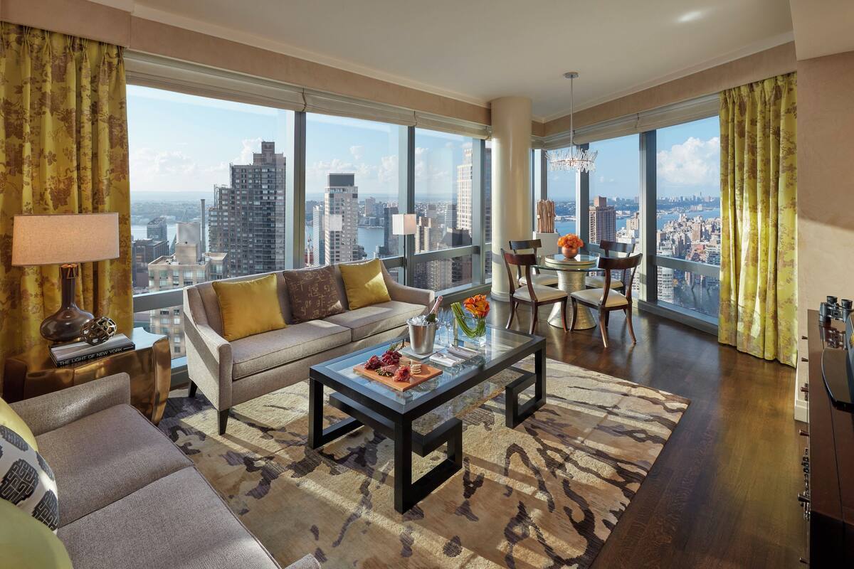 A modern living room with a sofa, coffee table, and dining area, featuring large windows with a city view.