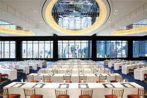 A spacious conference room with multiple tables and chairs, large windows offering a city view, and a prominent chandelier.
