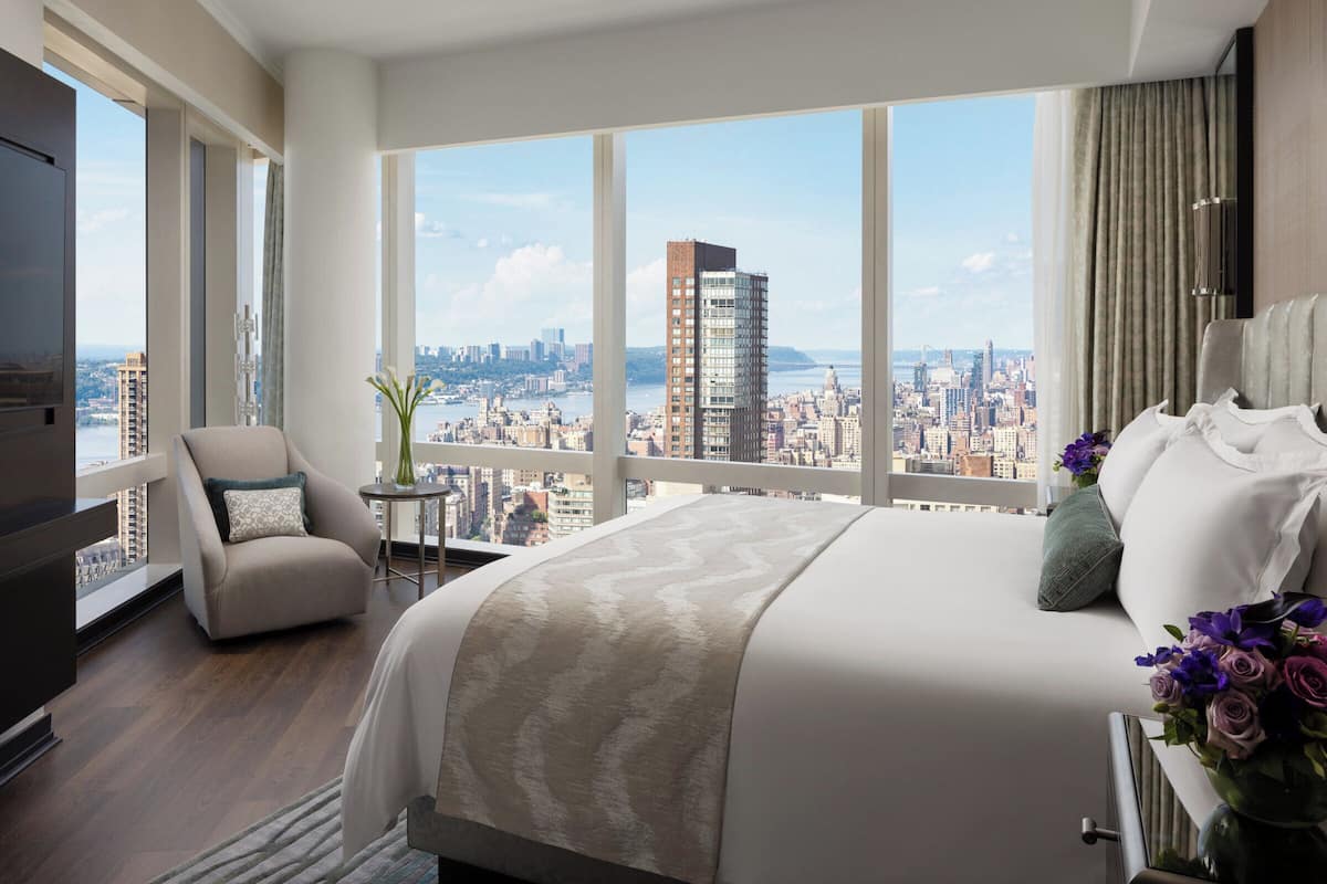 Een moderne hotelkamer met een groot bed, een stoel, een tafeltje met een vaas en uitzicht op de stad door grote ramen.