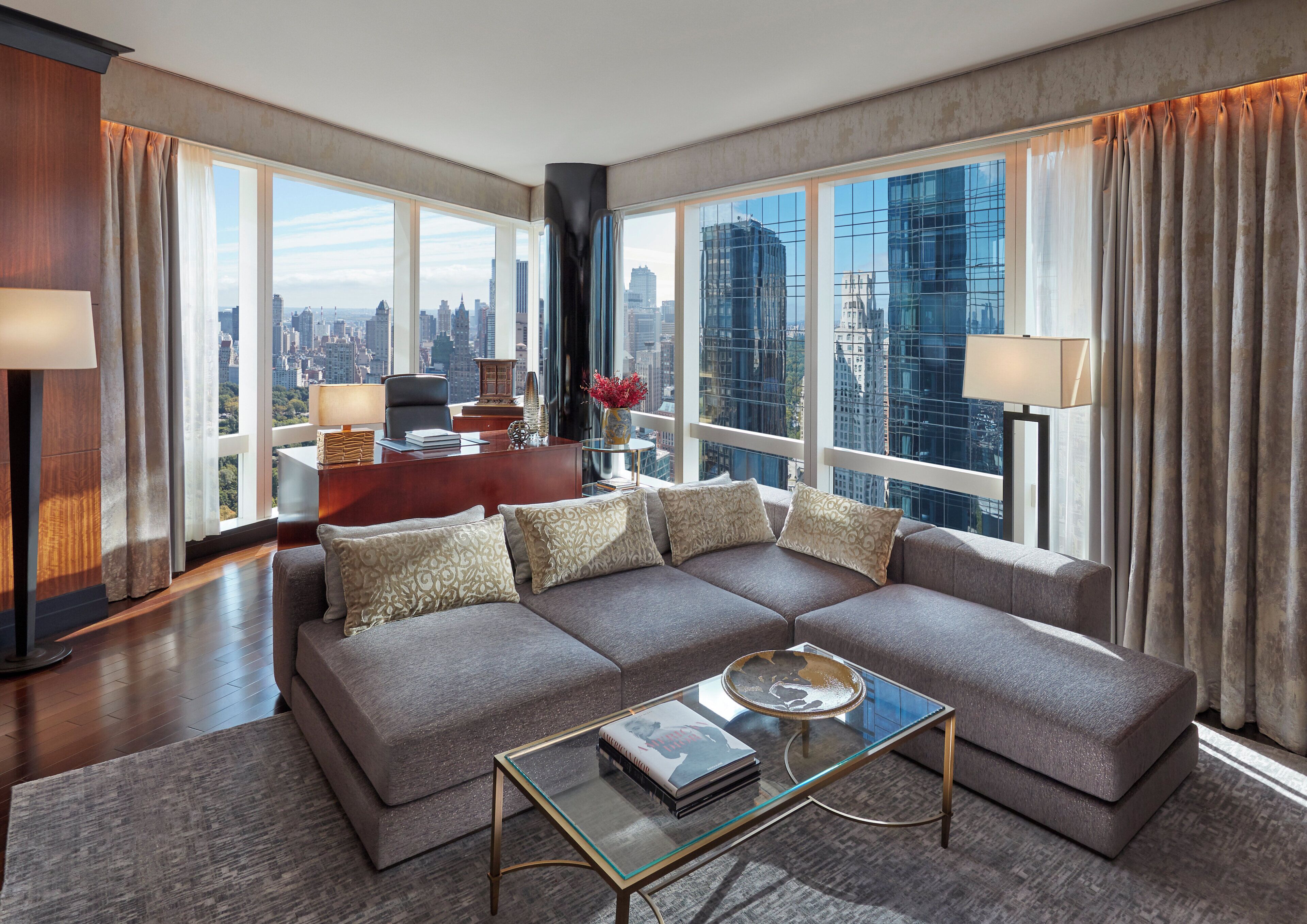 Un salon moderne avec un grand canapé d’angle, une table basse en verre et un bureau avec une chaise. De grandes fenêtres offrent une vue sur le paysage urbain.