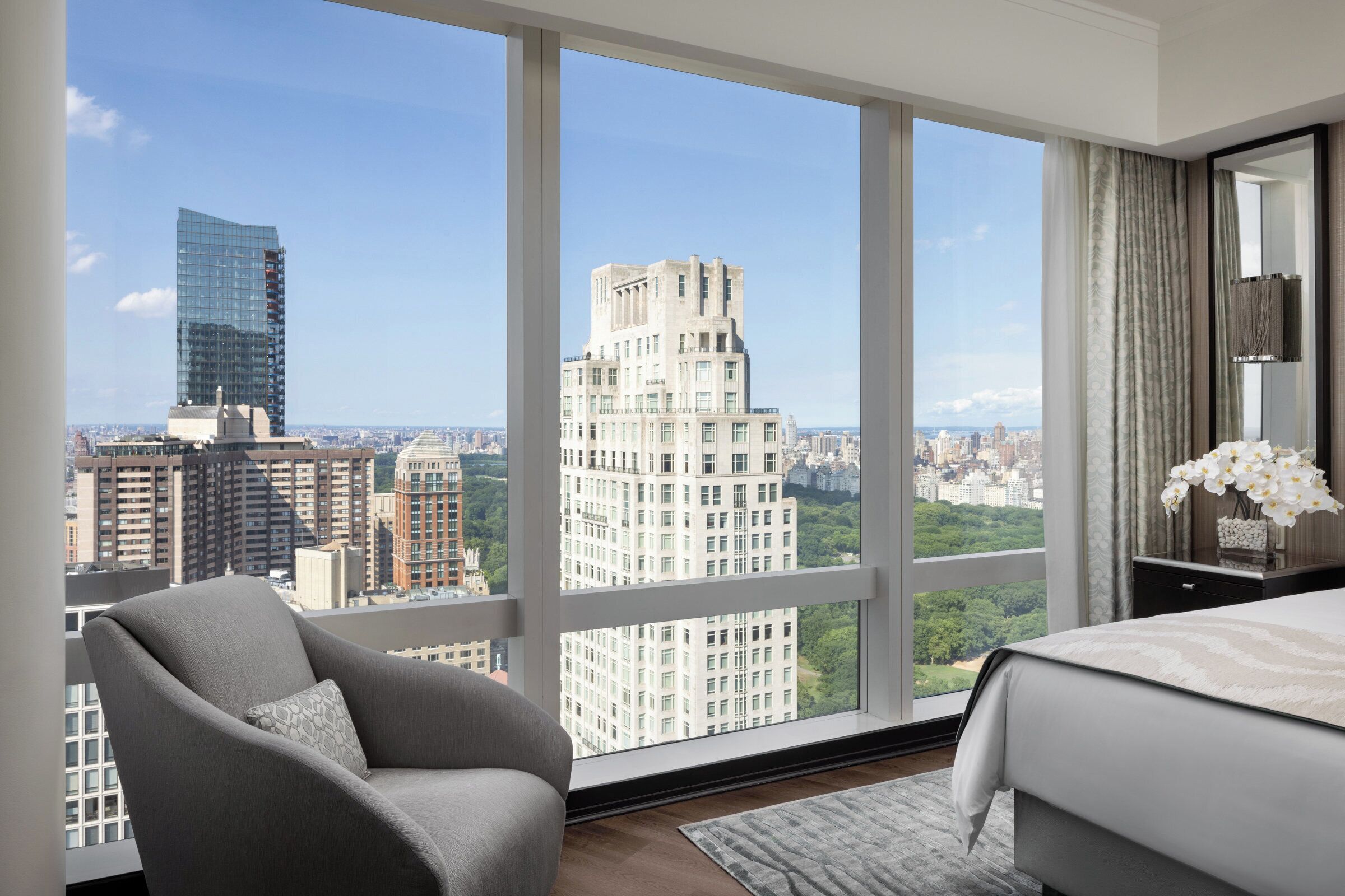 A modern hotel room with a large window offering a city view, a comfortable chair, a neatly made bed, and a small bedside table with a lamp.