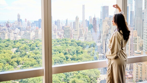 A person standing by a large window overlooking a cityscape with dense urban buildings and a green park area.