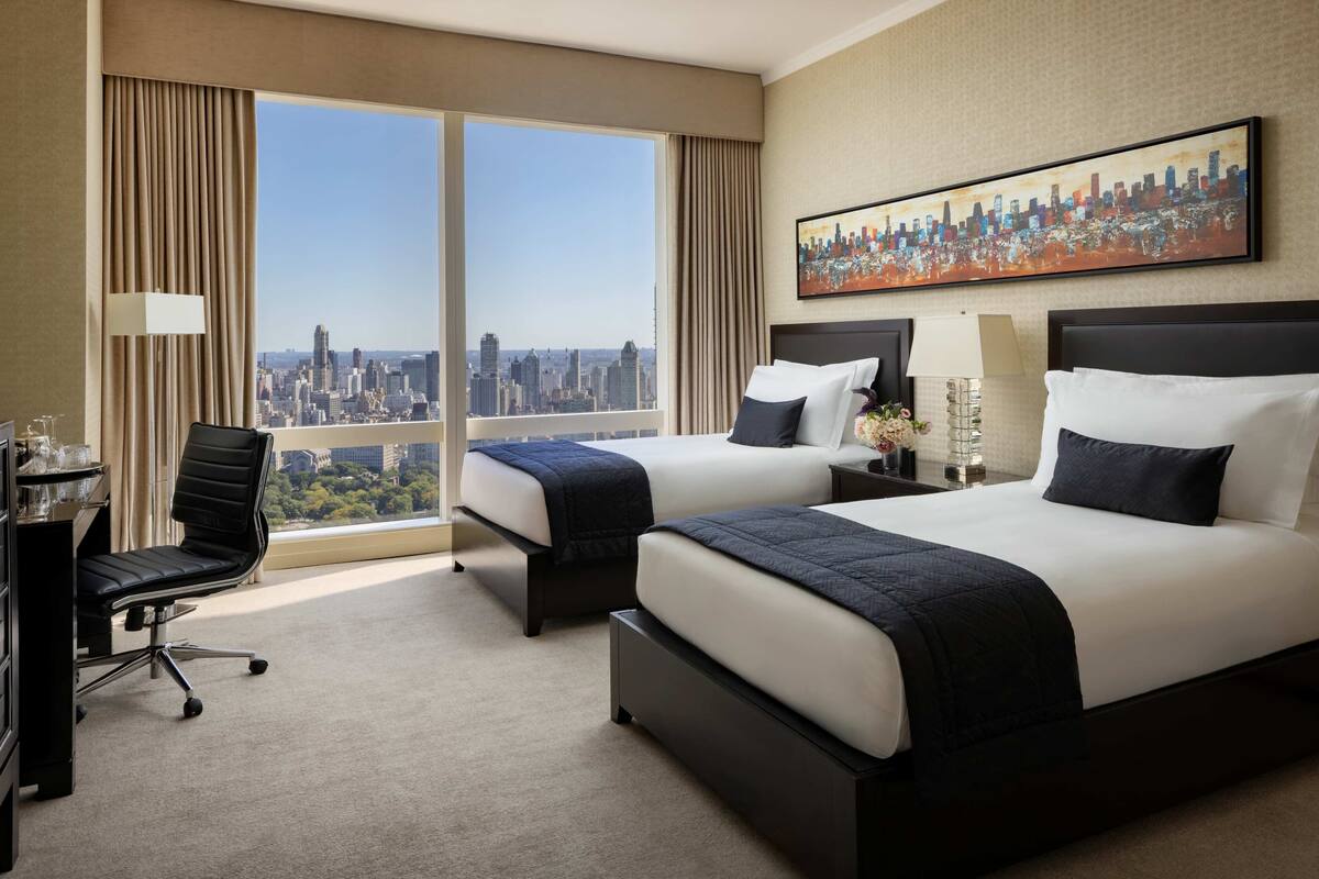 A hotel room with two beds, a desk, a chair, and a large window with a city view.