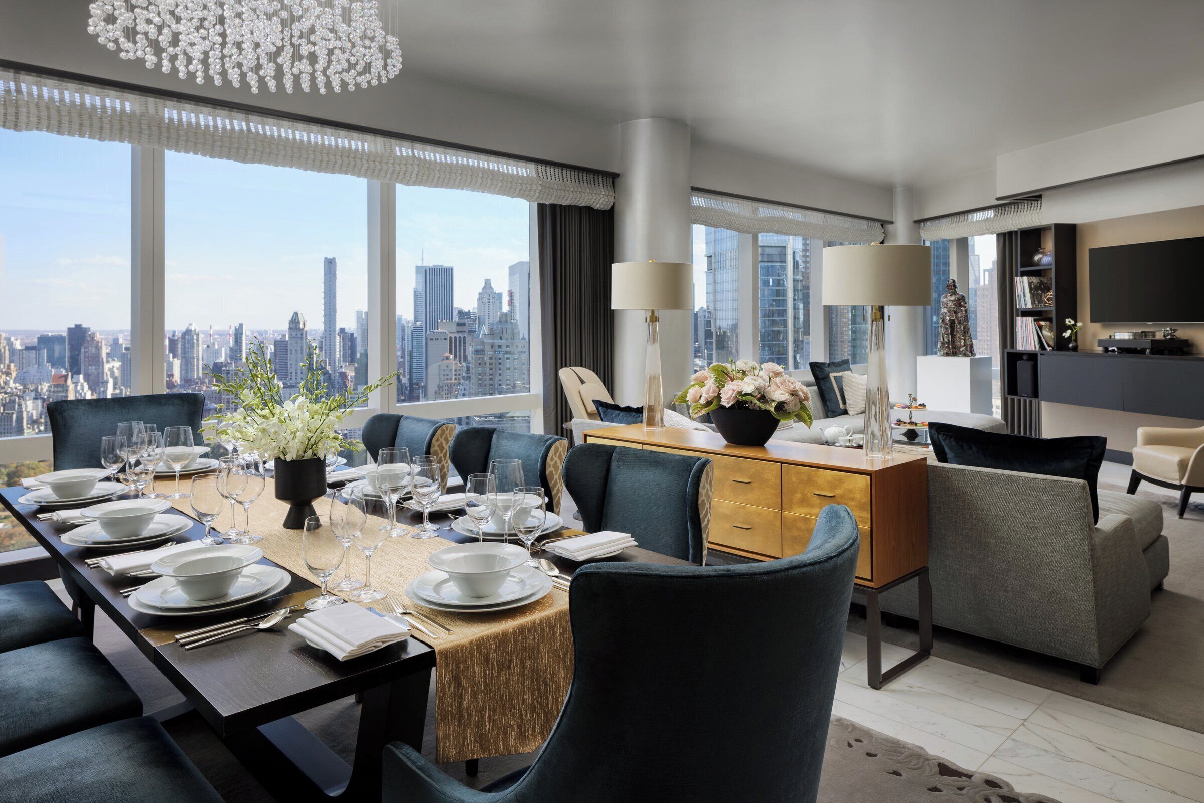 A modern hotel dining area with a city view, a long table set for dining, and a cozy seating area with a flat-screen TV.