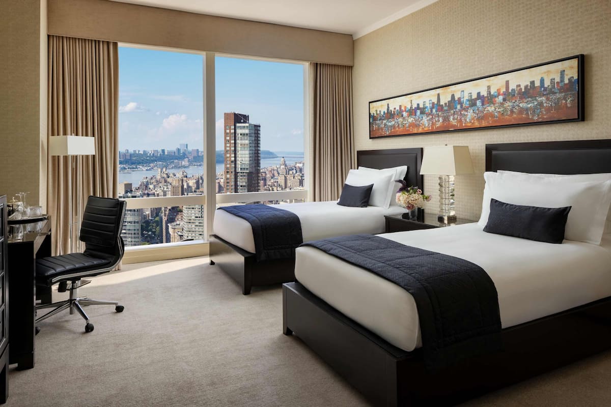 Een hotelkamer met twee bedden, een bureau, een stoel en een groot raam met uitzicht op de stad.