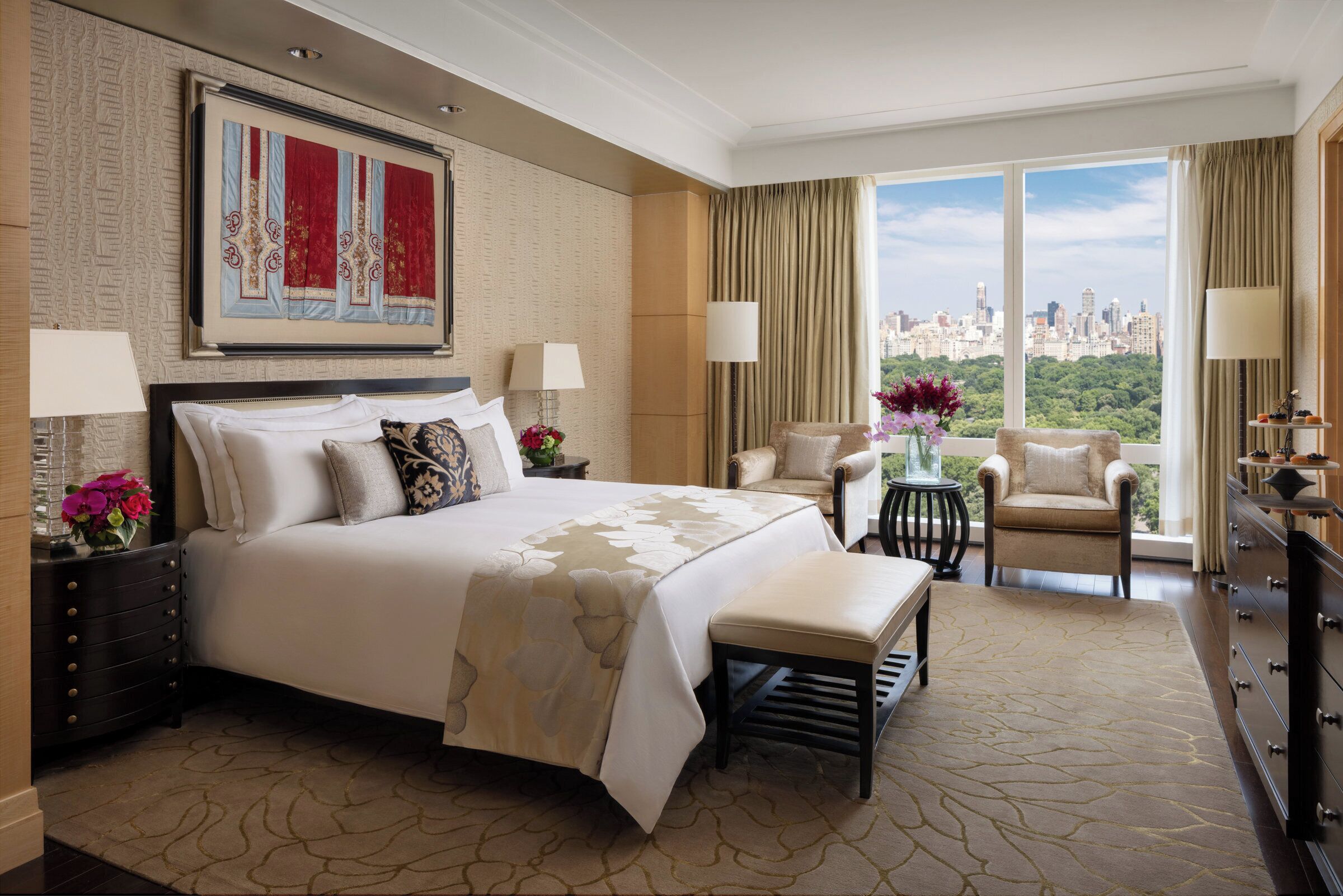 Ruime hotelkamer met een groot bed, twee fauteuils, een klein tafeltje met bloemen en uitzicht op de stad door grote ramen.