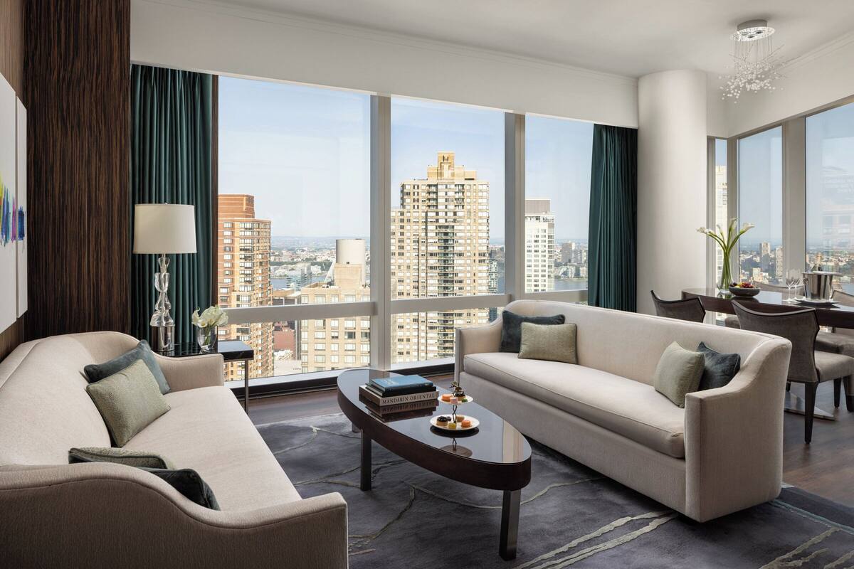 A modern hotel room with a large window offering a city view, a sofa, a coffee table, and a dining area.
