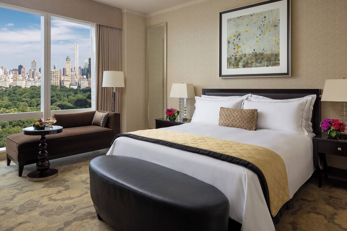 A hotel room with a large bed, a sofa, a round table, and a view of a cityscape.