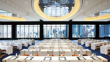 A spacious conference room with multiple tables and chairs, large windows offering a city view, and a prominent chandelier.