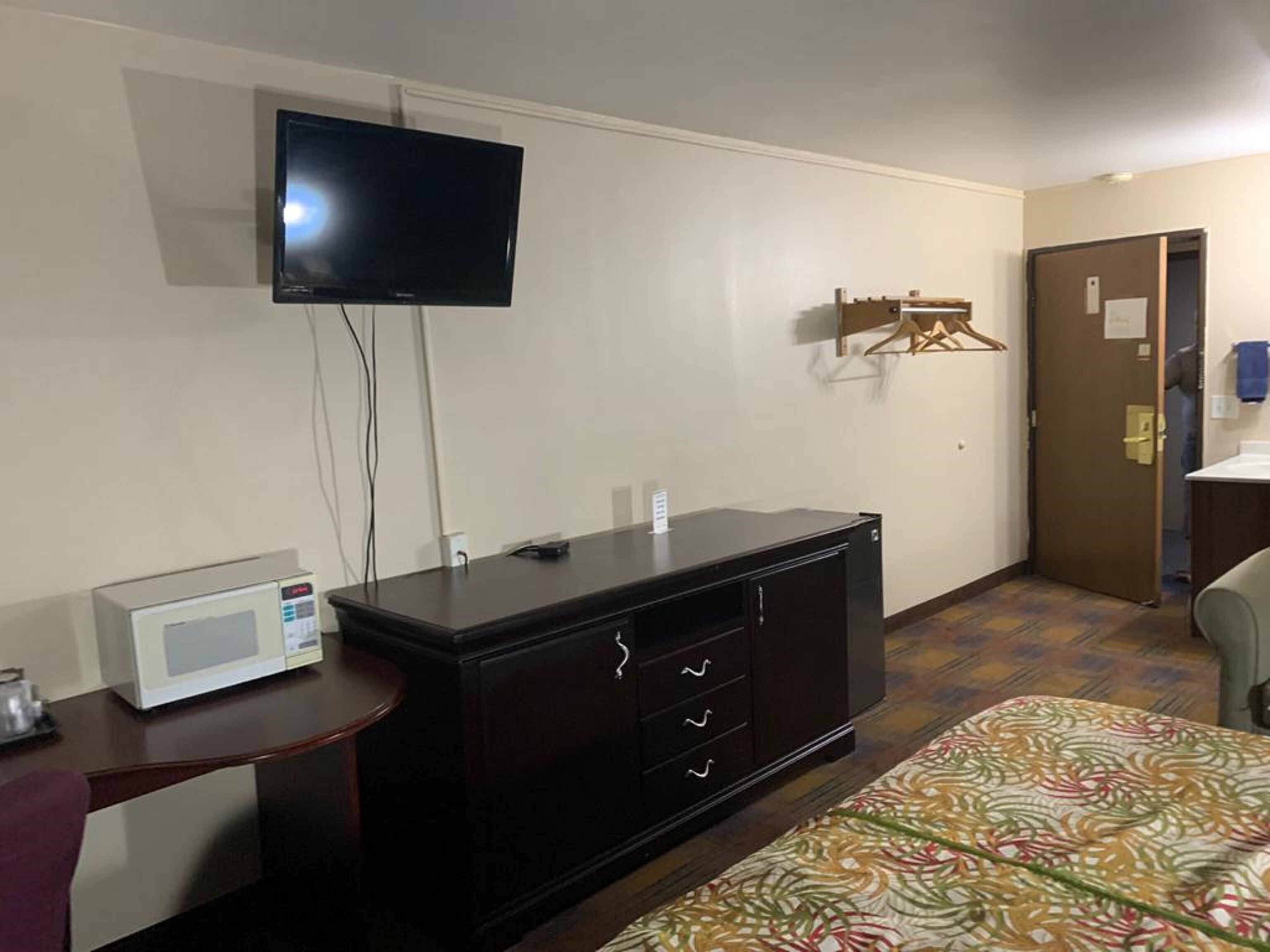 Une chambre d’hôtel dotée d’un téléviseur à écran plat fixé au mur, d’une commode en bois foncé avec des tiroirs, d’un four à micro-ondes posé sur une petite table, d’une couvre-lit à motifs et d’une porte donnant accès à la salle de bain.