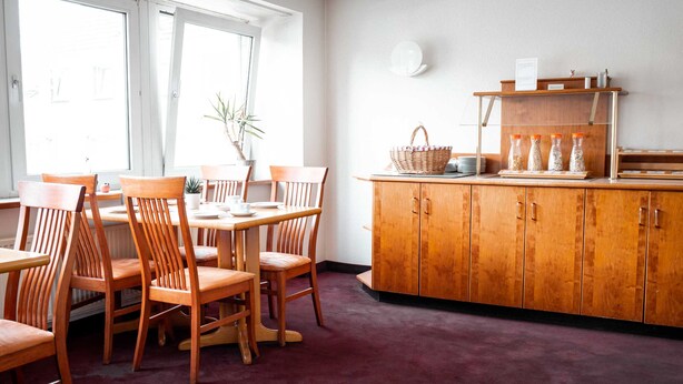 A dining area with wooden chairs and a table, a wooden cabinet with shelves, and a window with a plant.