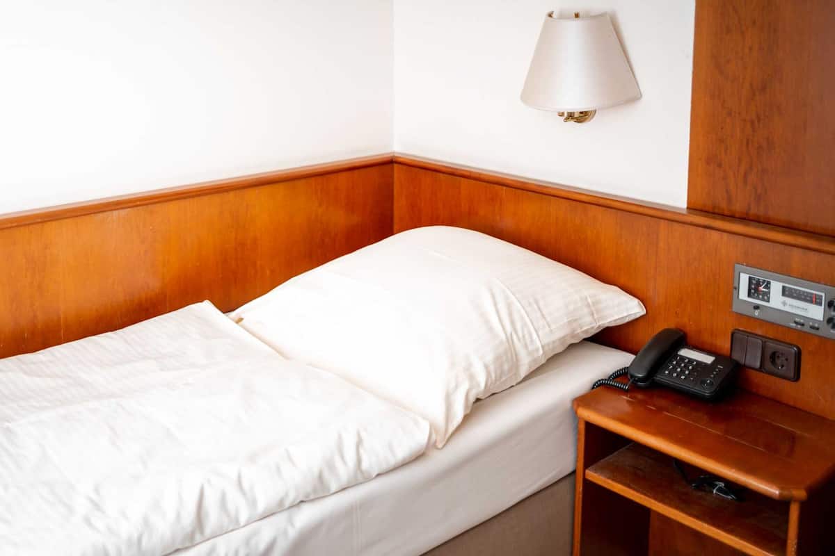 A single bed with white bedding, a bedside telephone, and a wall-mounted lamp.