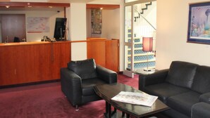 A hotel lobby with a reception desk, two black leather sofas, a small table with papers, and a staircase leading to an upper floor.