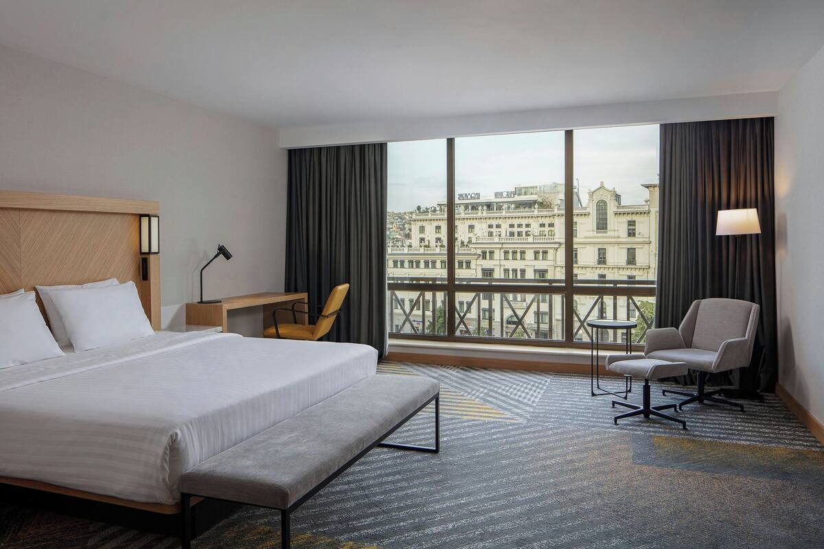 A hotel room with a large bed, a desk, a chair, and a view of a cityscape through large windows.