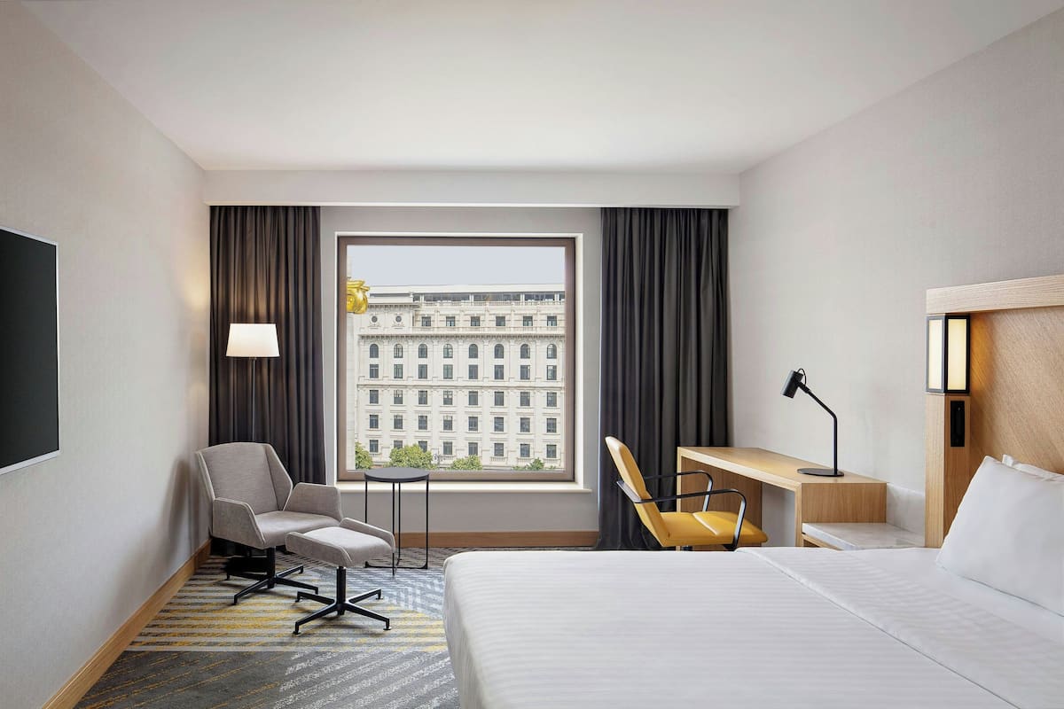 A hotel room with a bed, a desk, a chair, a lamp, and a view of a building through a window.