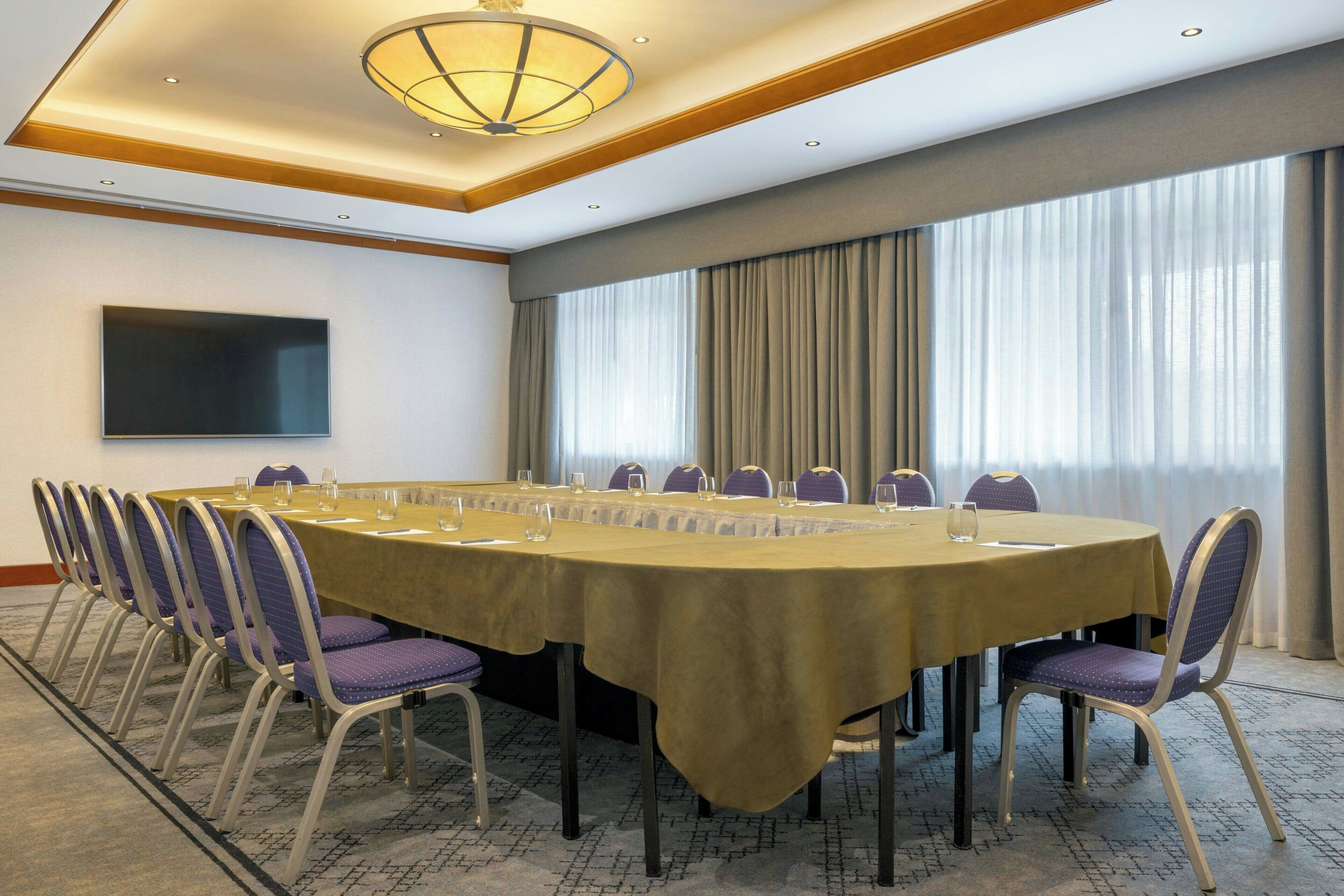 Une salle de conférence équipée d’une longue table, de chaises, d’un téléviseur à écran plat et de grandes fenêtres avec des rideaux.