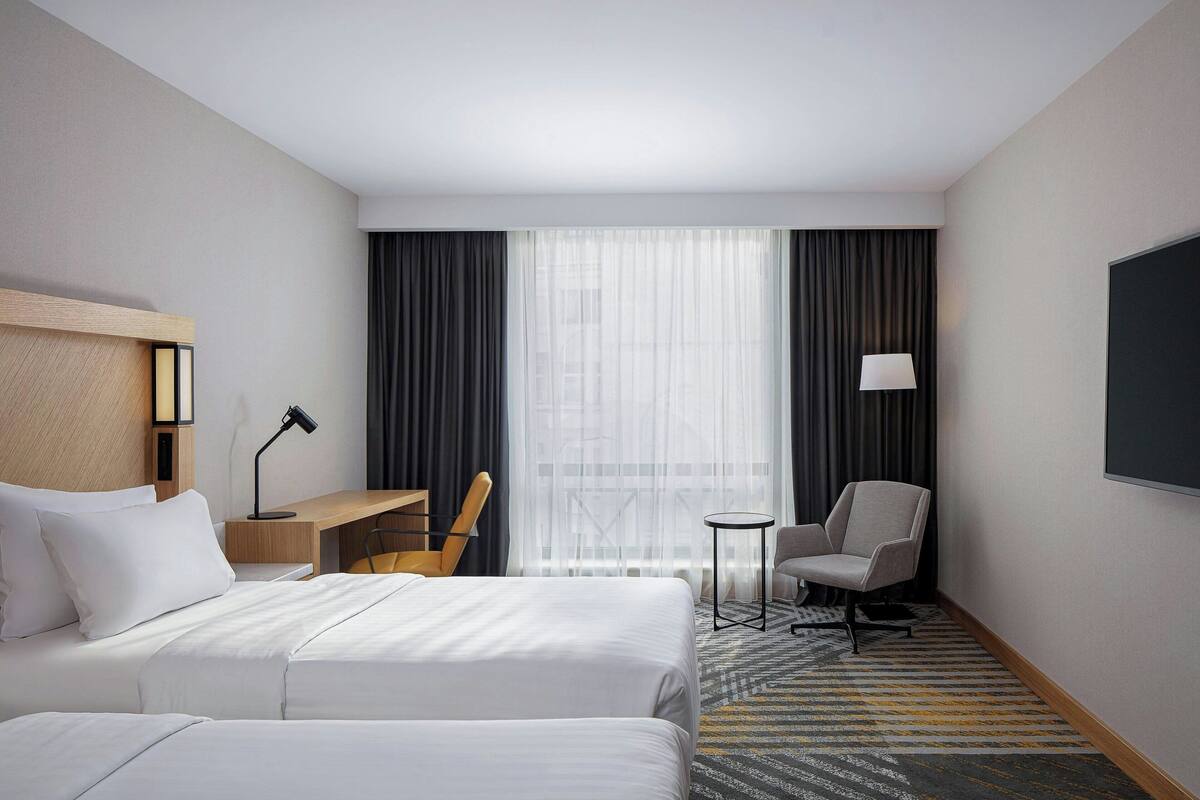 A hotel room with two beds, a desk, a chair, a TV, and a large window with curtains.