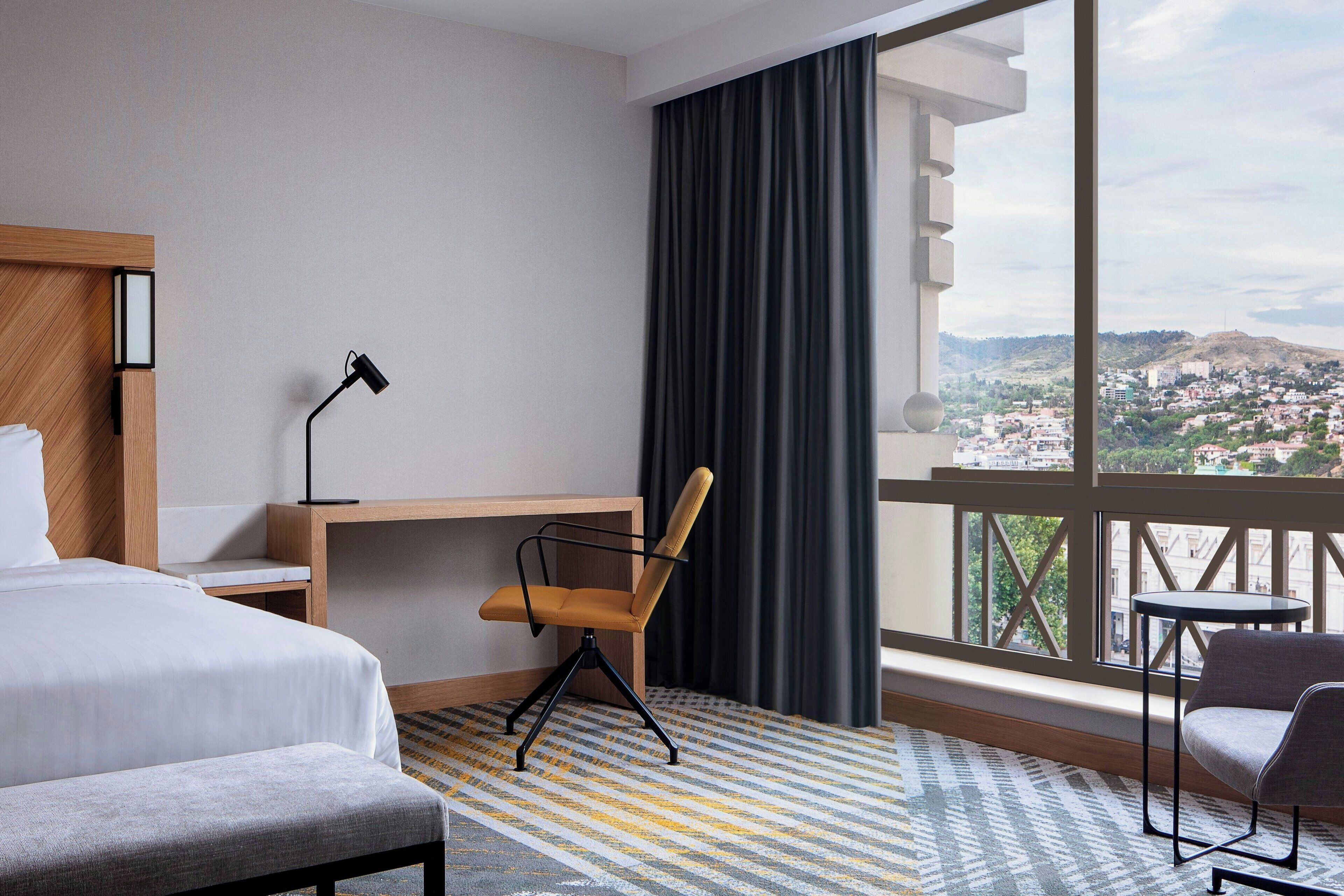 A hotel room with a bed, a desk, a chair, and a view of a cityscape through large windows.
