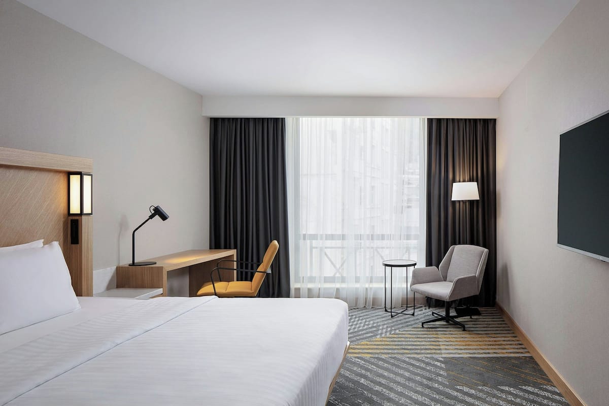 A hotel room with a large bed, a desk with a chair, a small table with a chair, and a large window with curtains.