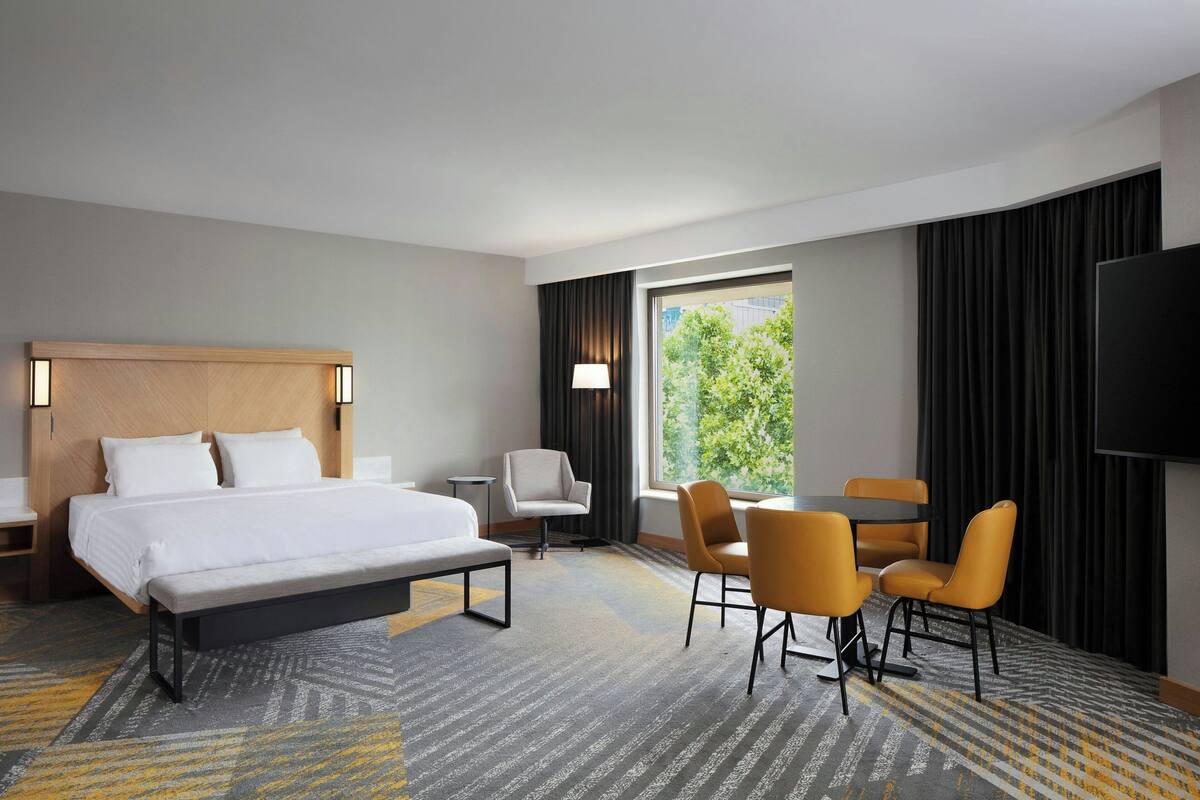 A hotel room with a bed, bedside tables, a desk, two chairs, a TV, and a large window with curtains.