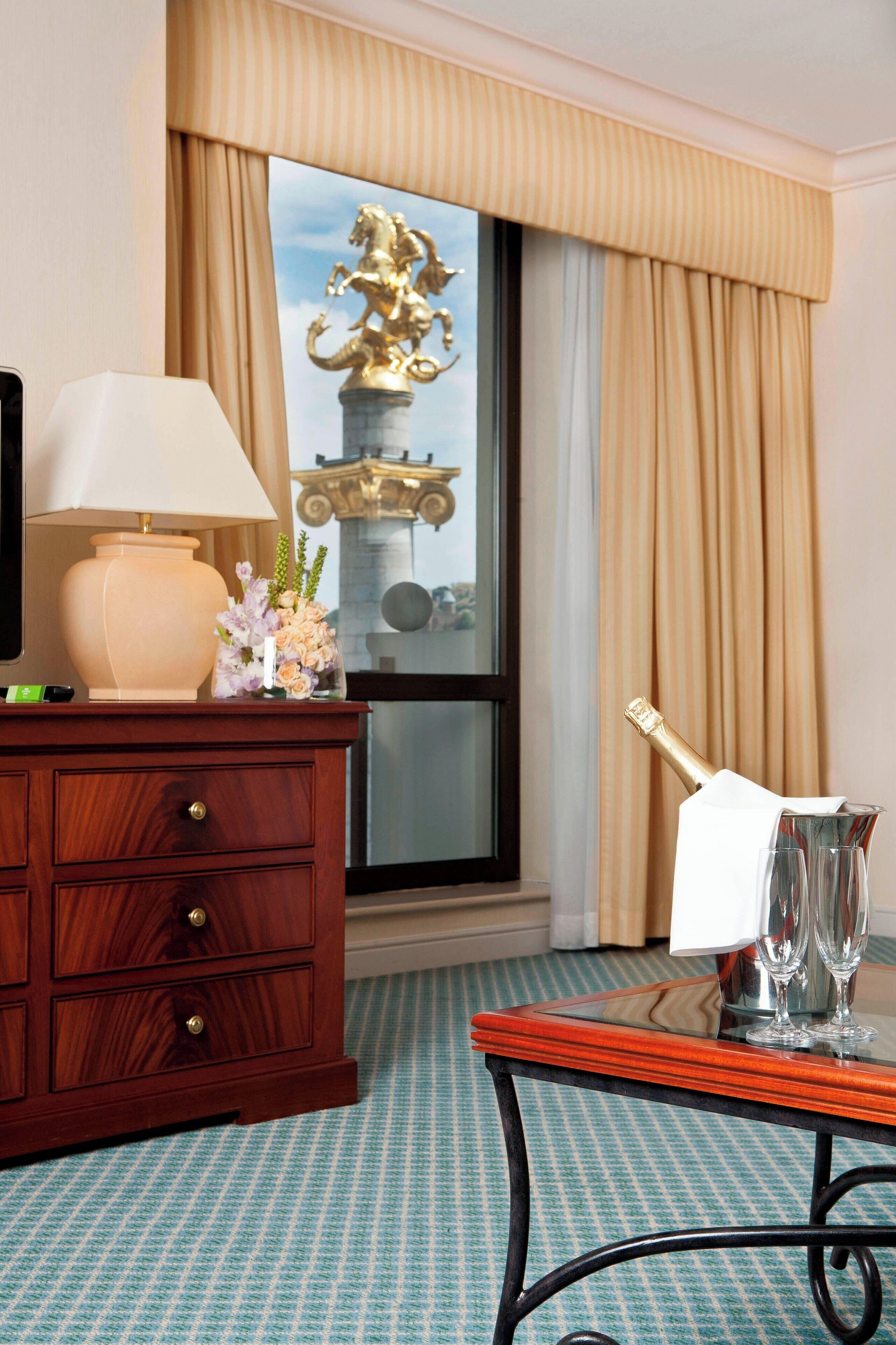 Habitación de hotel con cómoda de madera, lámpara, florero con flores, botella de champán y vista a una estatua a través de una ventana.