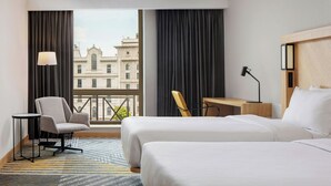 Habitación de hotel con dos camas, un escritorio, una silla, una lámpara y vistas a un edificio a través de ventanas grandes.