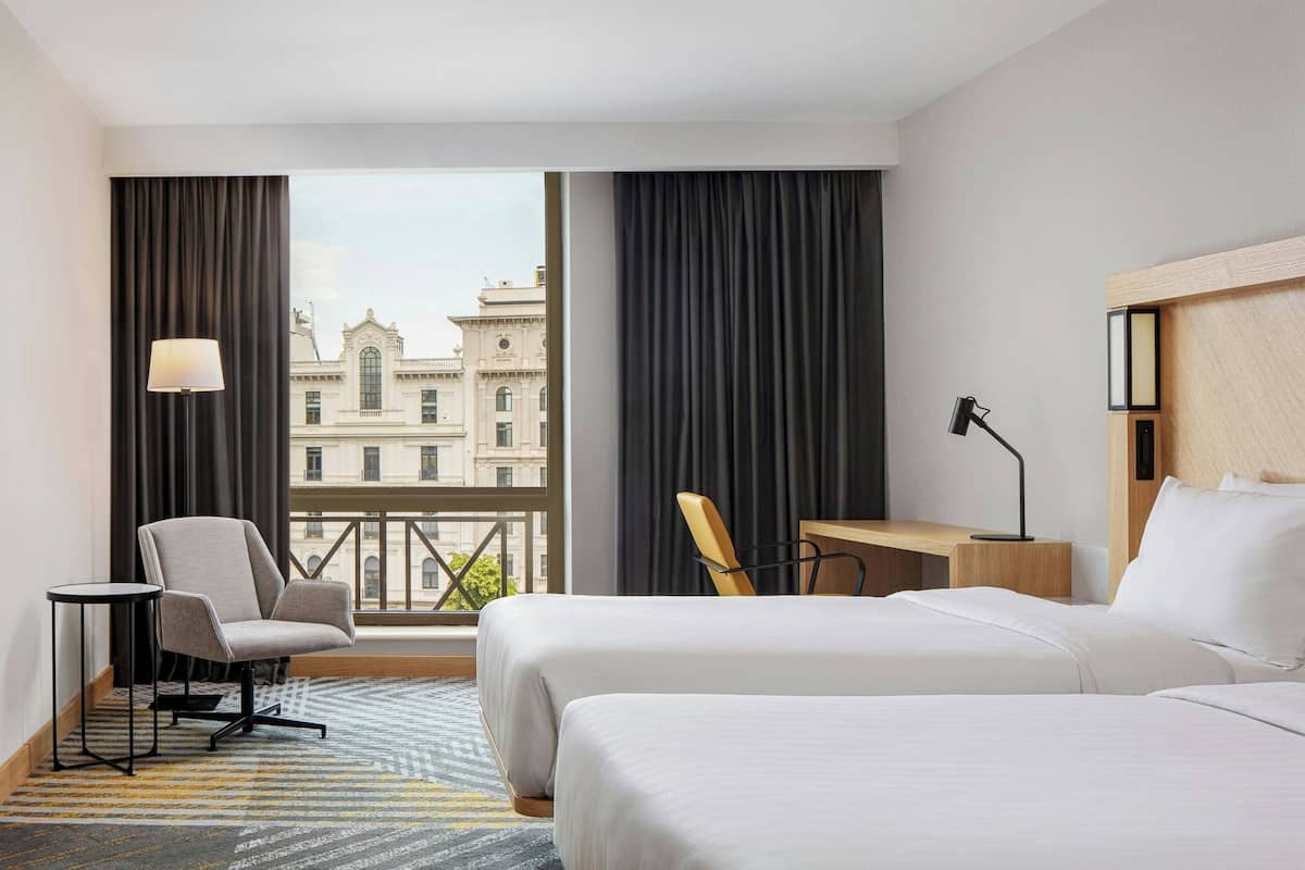 A hotel room with two beds, a desk, a chair, a lamp, and a view of a building through large windows.