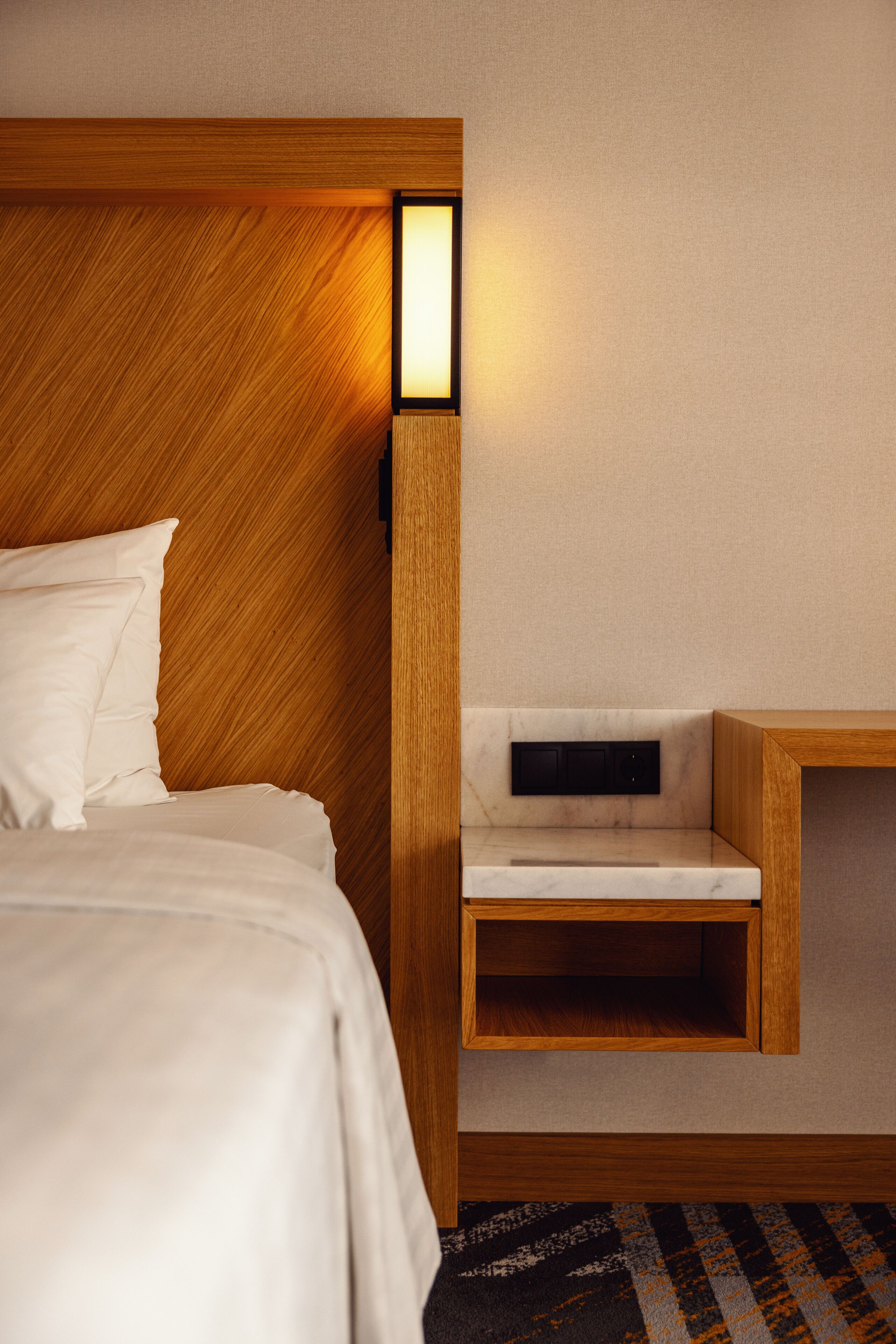 A modern hotel room with a wooden headboard, a bedside table with a built-in lamp, and a small wooden nightstand.