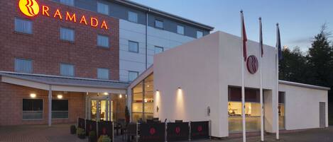 A Ramada hotel with a modern facade, outdoor seating area, and flags.