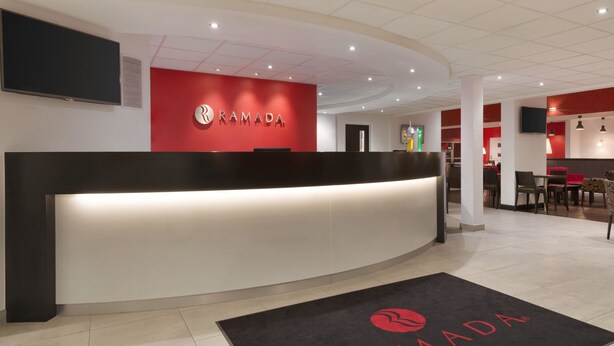 A reception area with a curved desk, a red wall with the 'RAMADA' logo, and a black doormat with the same logo. - Ramada by Wyndham Wakefield (Ossett)