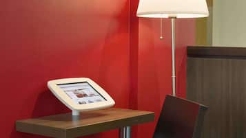 A modern hotel reception area with a red wall, a digital information kiosk, and a floor lamp.