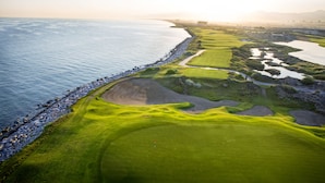 A coastal golf course with a sandy green, water hazards, and a rocky shoreline.