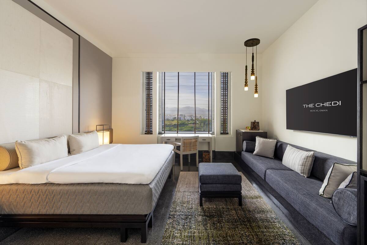 A modern hotel room with a large bed, a sofa, a desk, and a view of the city.