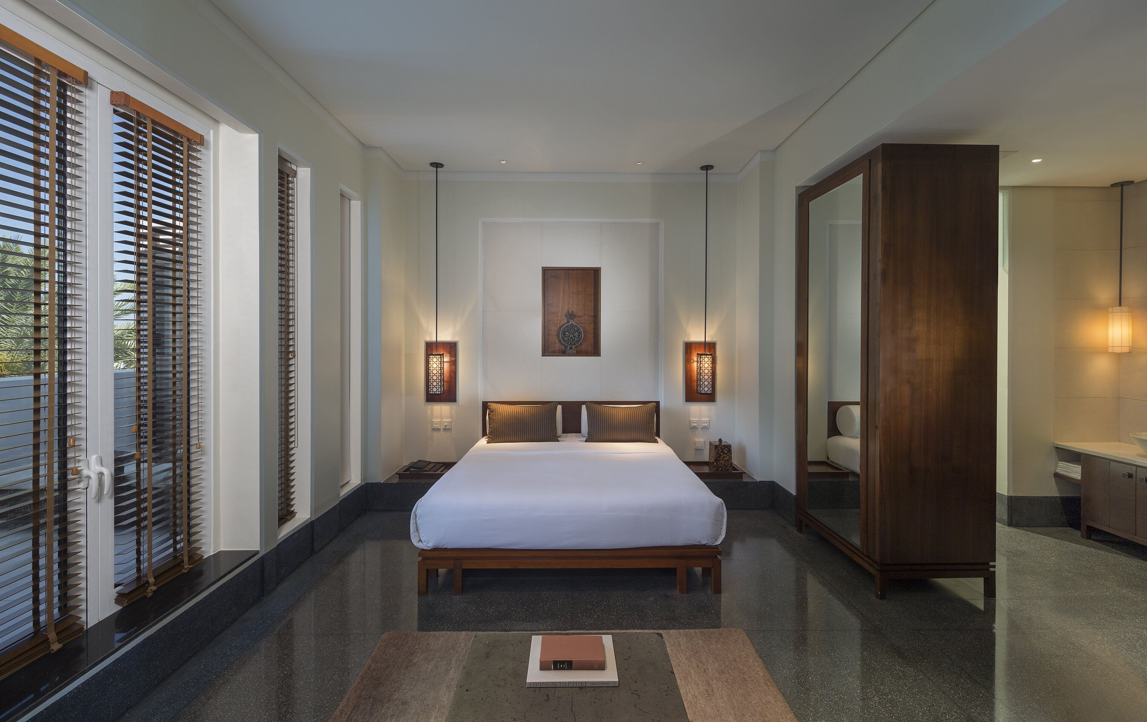 A spacious bedroom with a large bed, wooden blinds, a wall-mounted light fixture, and a mirror.