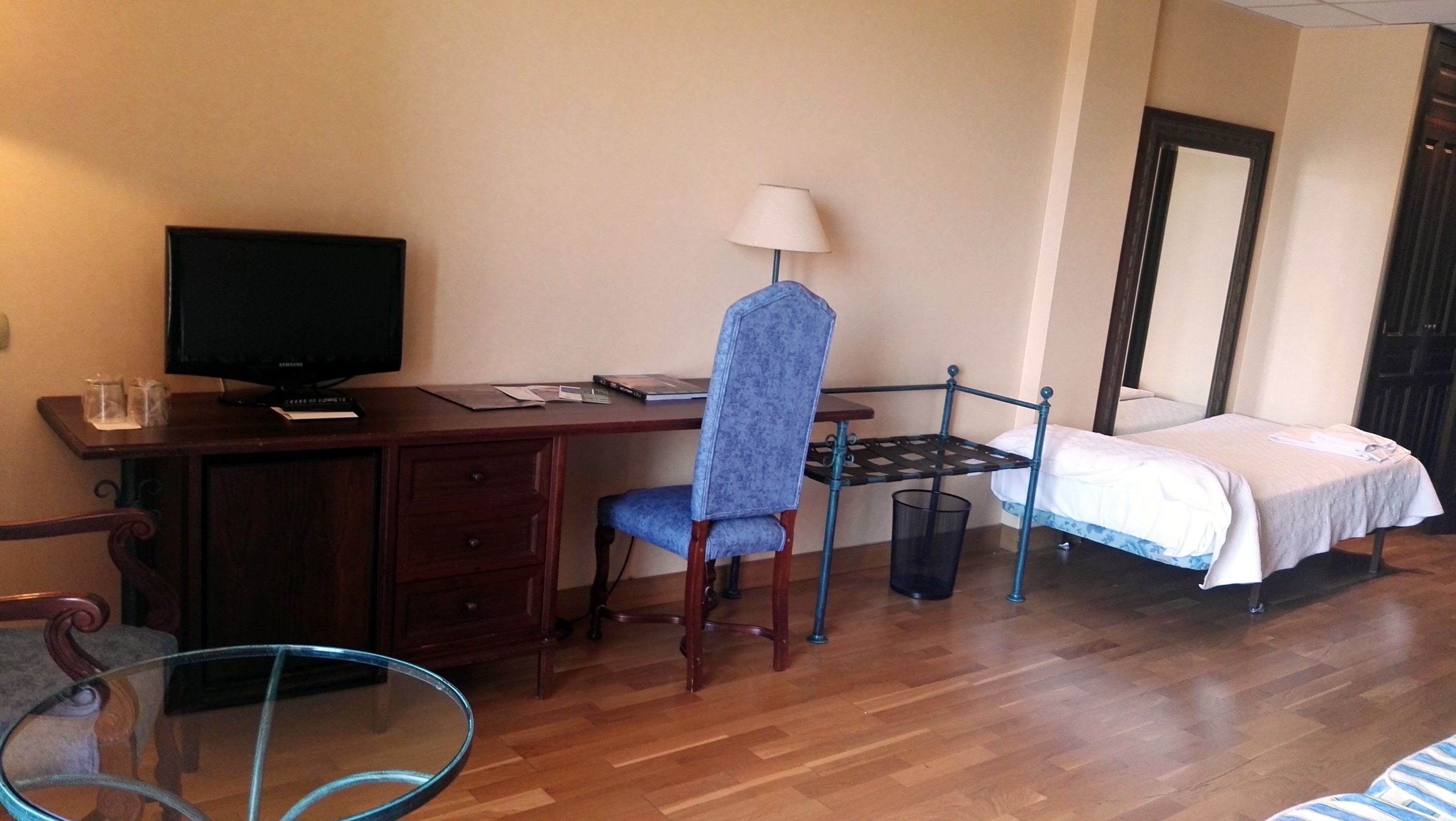 Hotellihuone, jossa on puinen työpöytä, tuoli, valkoisella päiväpeitteellä varustettu sänky, televisio ja pieni pöytä, jossa on lamppu.