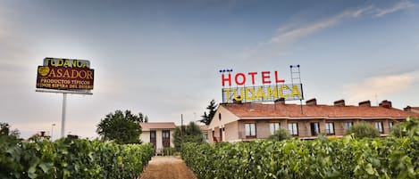 A vineyard with rows of grapevines leading to a building with a sign that reads "TUDANCA ASADOR" and "HOTEL TUDANCA".