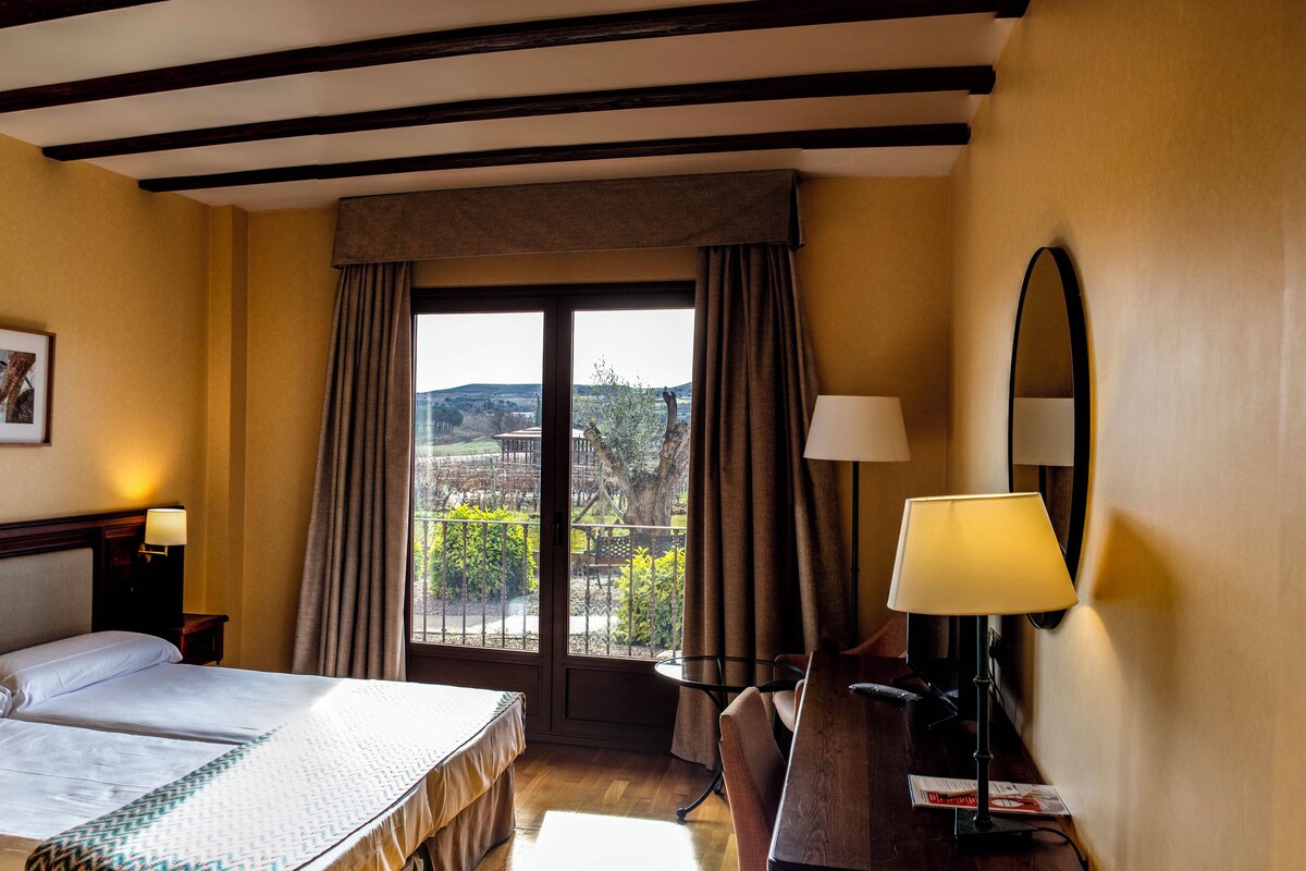 A hotel room with a double bed, a desk with a lamp, a chair, a window with curtains, and framed pictures on the wall.