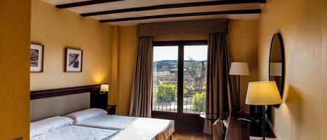 Una habitación de hotel con cama doble, un escritorio con lámpara, una silla, una ventana con cortinas y cuadros en la pared.