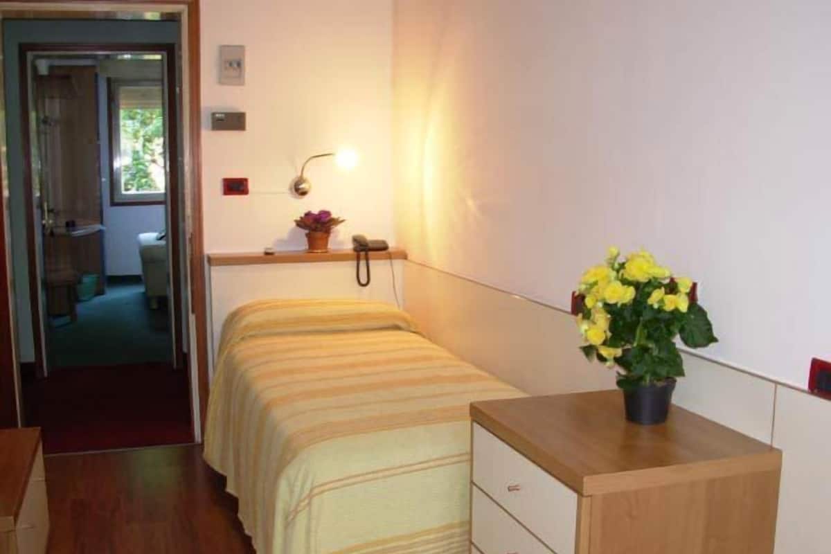 Ein kleines, ordentlich eingerichtetes Schlafzimmer mit einem Einzelbett, einem Nachttisch mit Blumenvase, einer Wandlampe und einer Holzkommode.