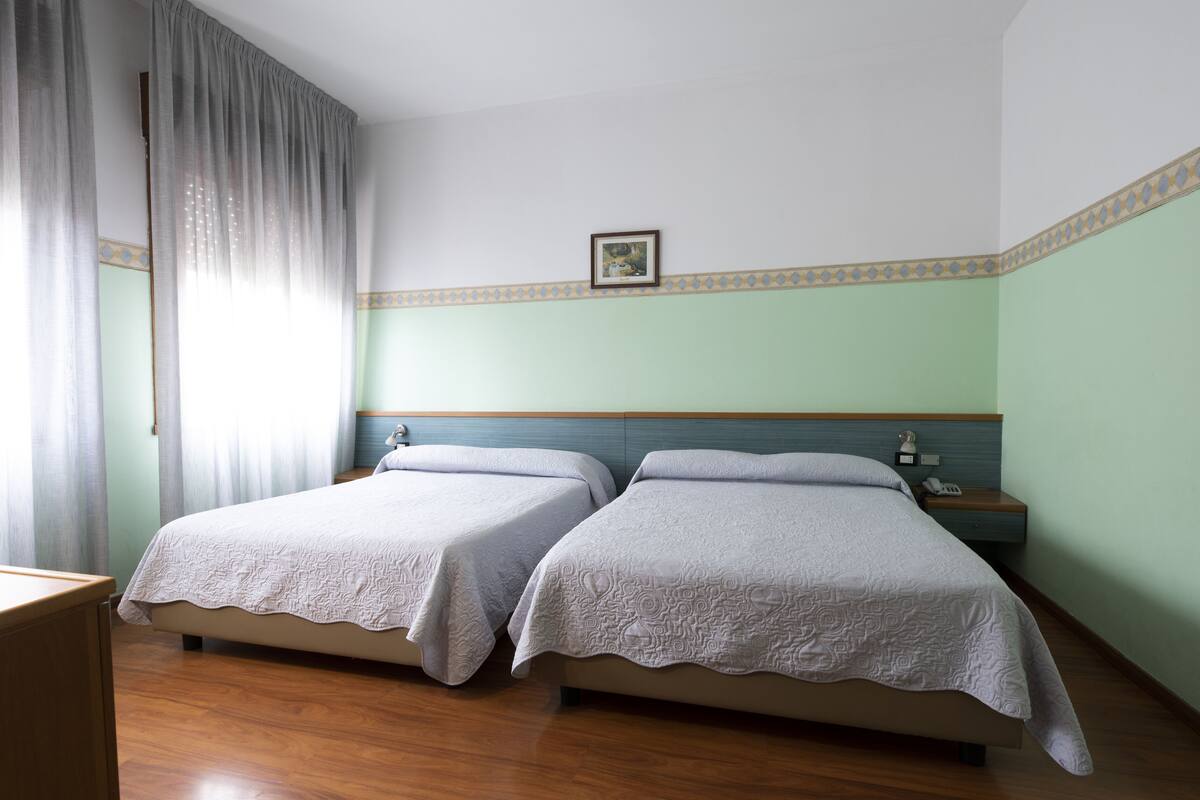 Ein Hotelzimmer mit zwei Betten, einem Nachttisch, einem gerahmten Bild an der Wand und einem Fenster mit Vorhängen.