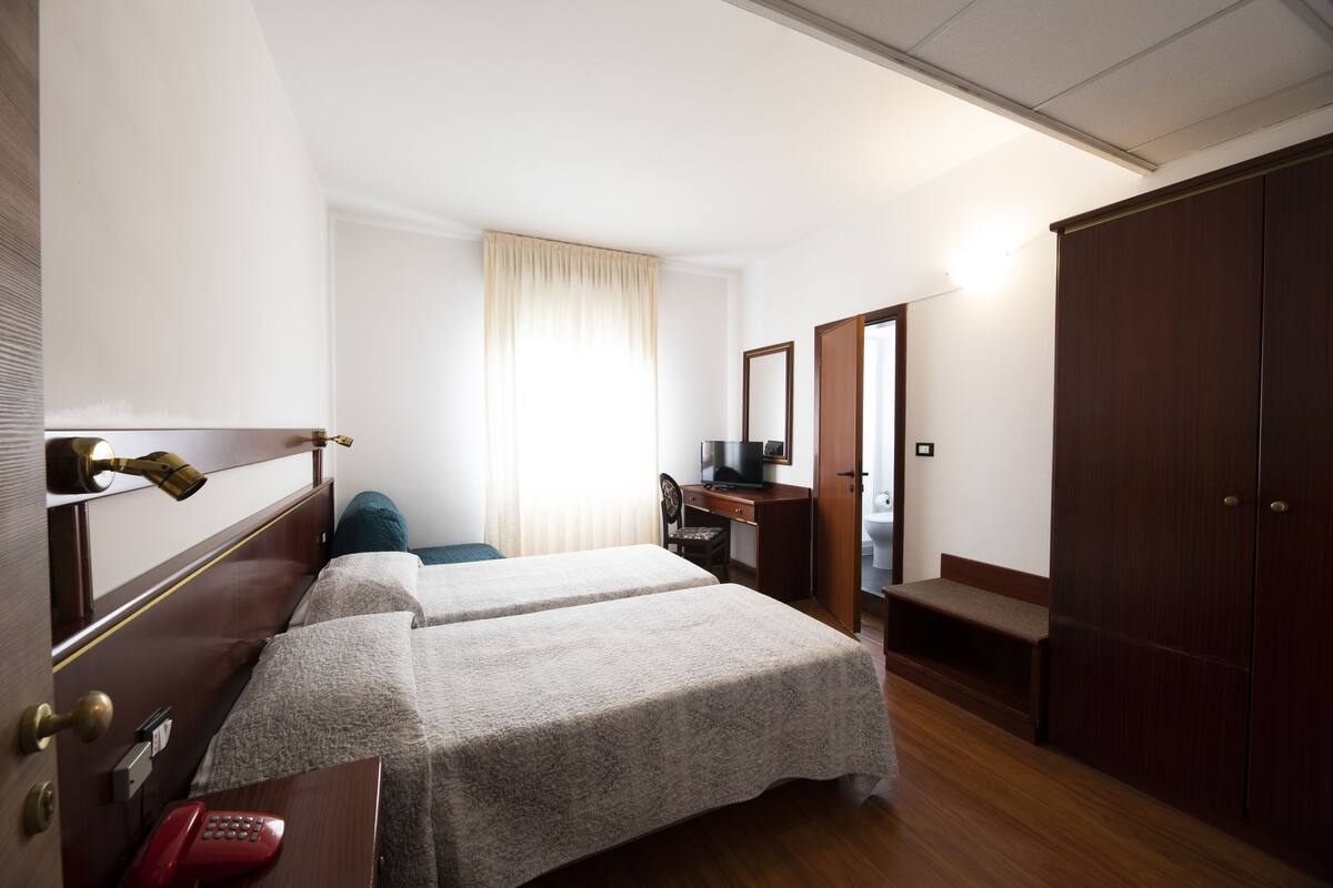 Ein Hotelzimmer mit zwei Betten, einem Schreibtisch, einem Stuhl, einem Kleiderschrank und einem durchgehenden Türspalt sichtbaren Badezimmer.