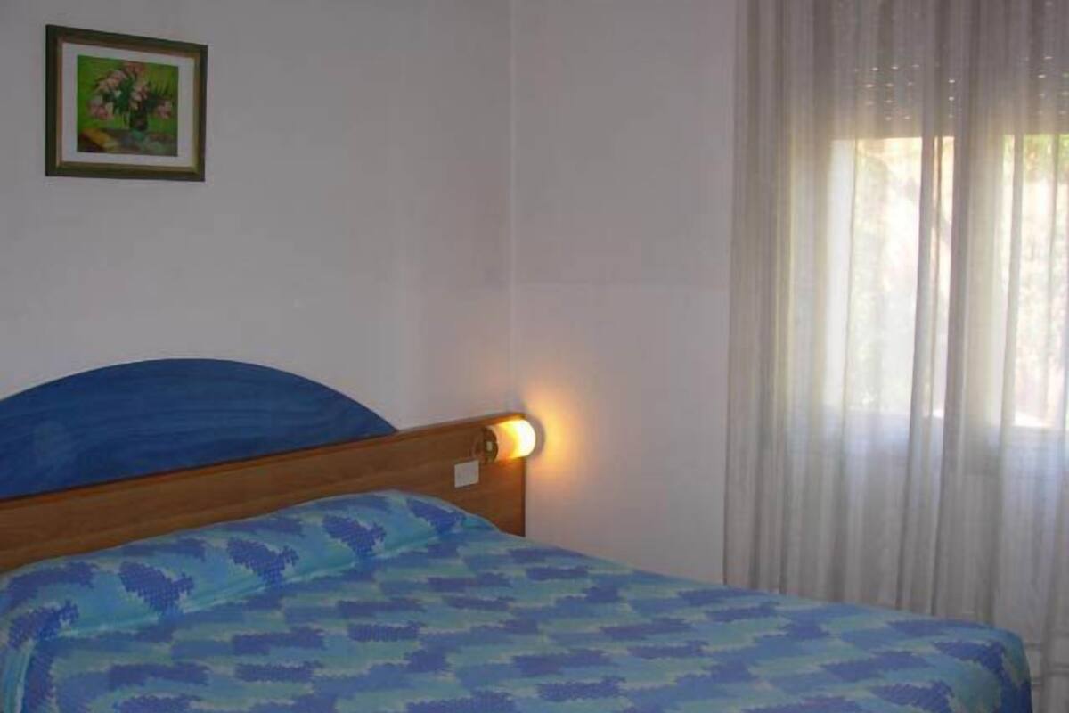 Ein Zimmer mit einem Bett, einem Nachttisch, einem gerahmten Bild an der Wand und einem Fenster mit Vorhängen.