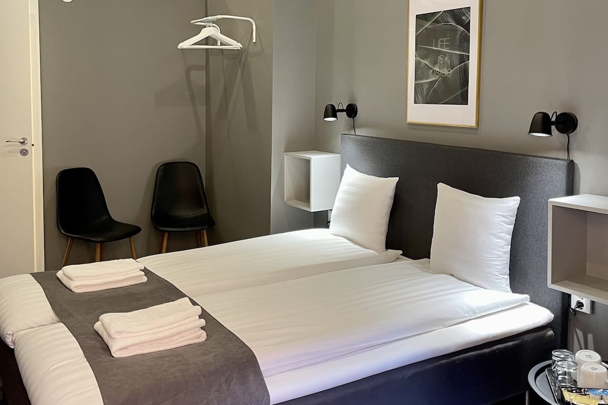 A modern hotel room with a large bed, two bedside tables, a headboard, a framed artwork, a wardrobe, and two chairs.