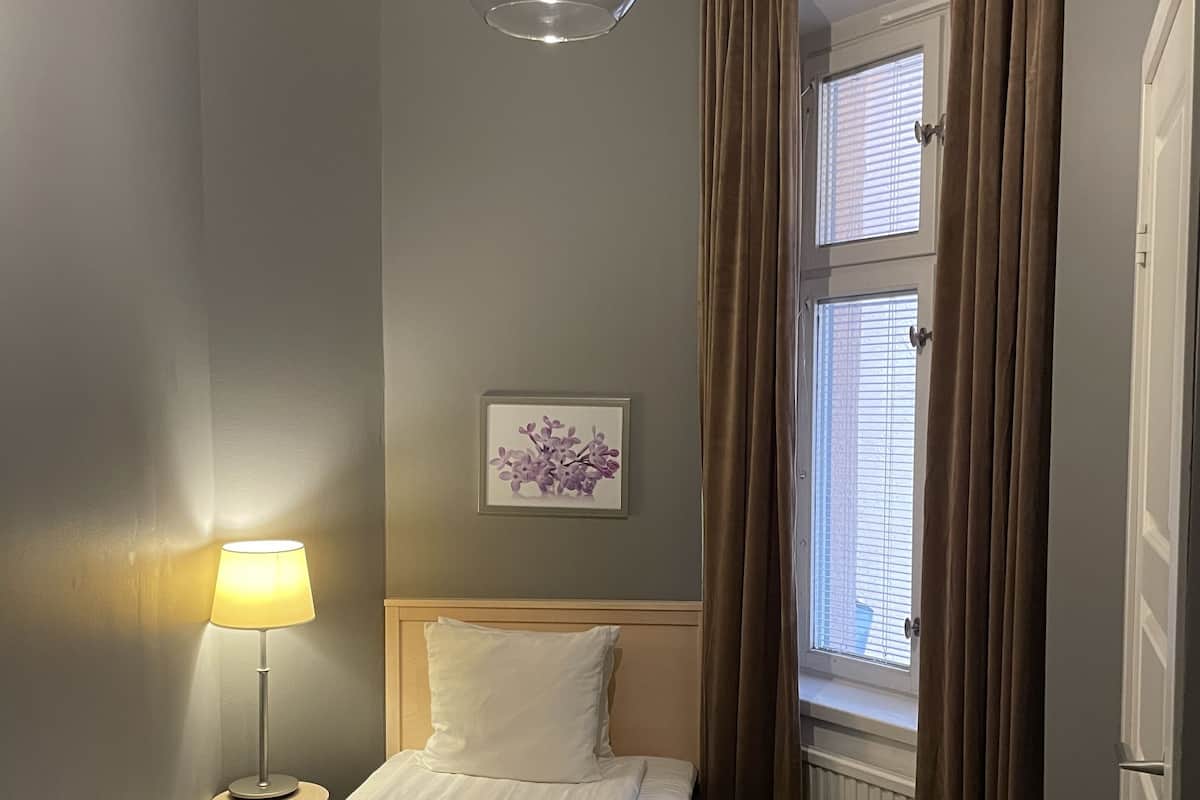 A neatly made bed with a pillow and a folded towel, a bedside lamp, a framed picture on the wall, and a window with curtains.