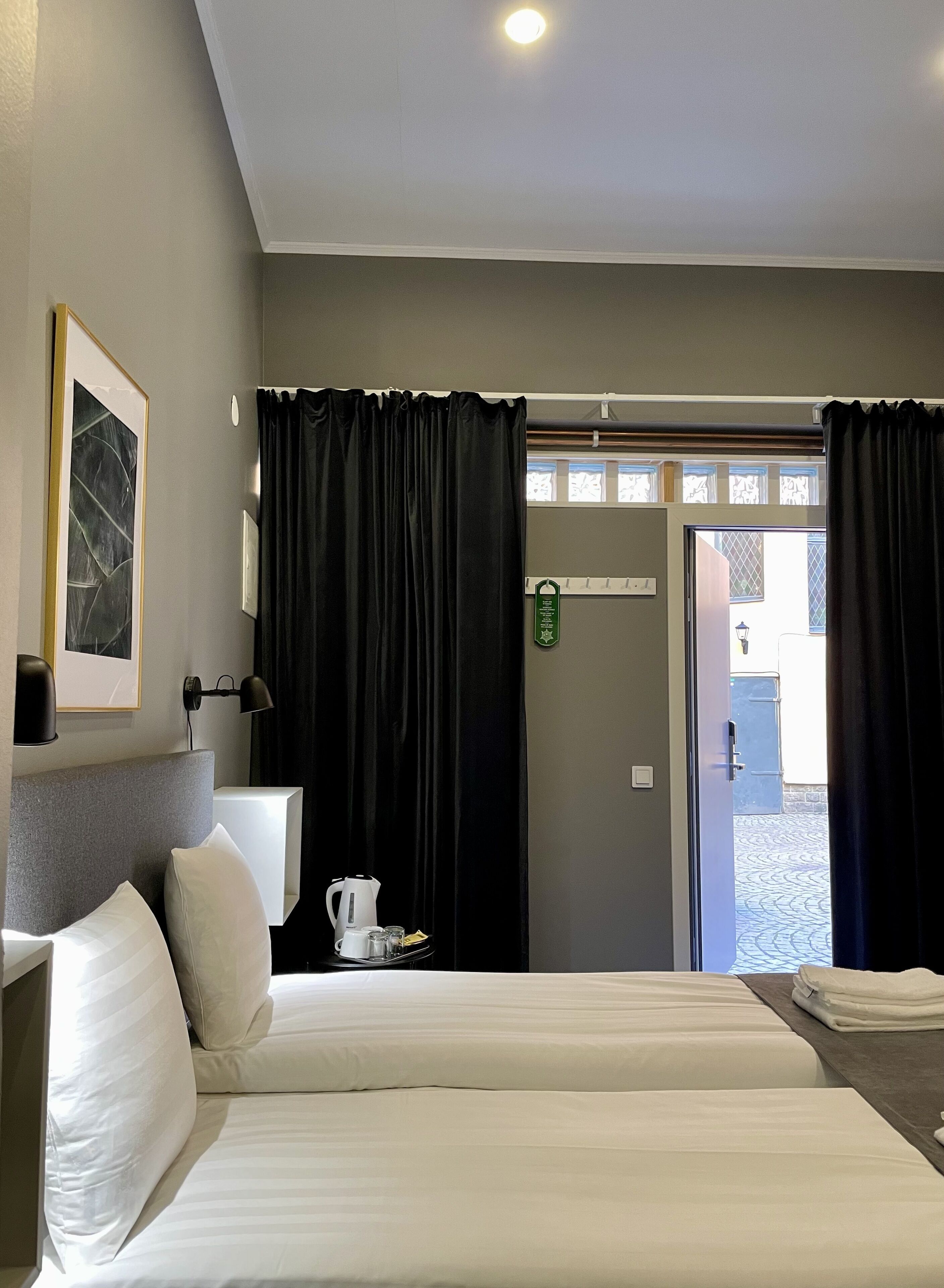 A hotel room with two beds, a coffee maker, a bedside table, a framed picture, and a view through a door.