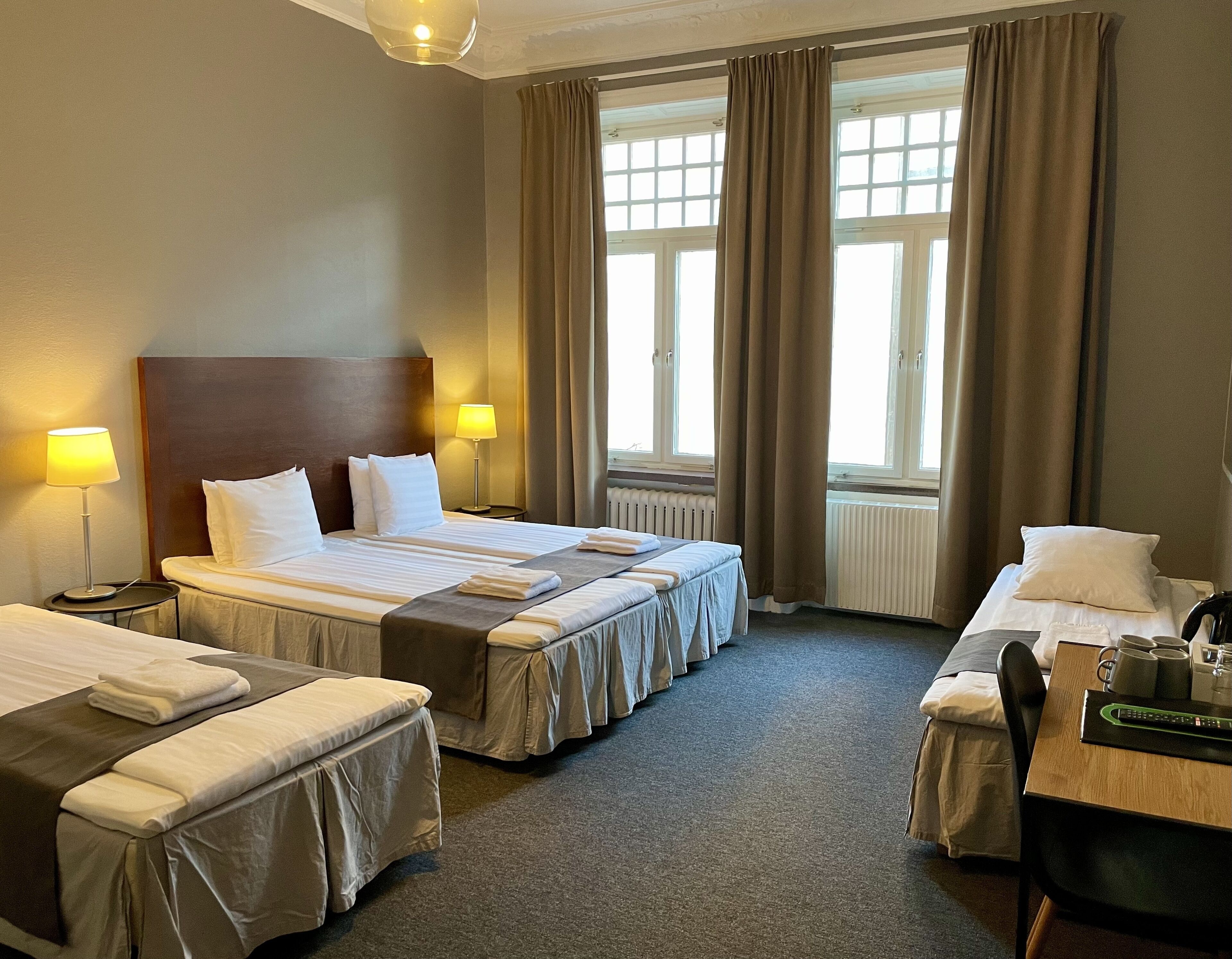 Une chambre d’hôtel avec deux lits, un bureau et une chaise. Il y a des lampes de chaque côté des lits et une fenêtre avec des rideaux.