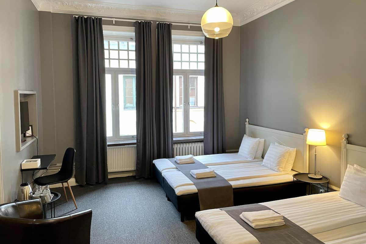 A hotel room with two beds, a desk, a chair, a lamp, and large windows with curtains.