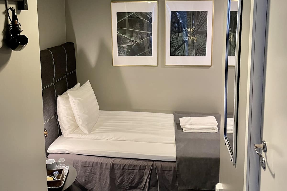 A compact hotel room with a single bed, a bedside table, a hanging coat rack, and two framed pictures on the wall.