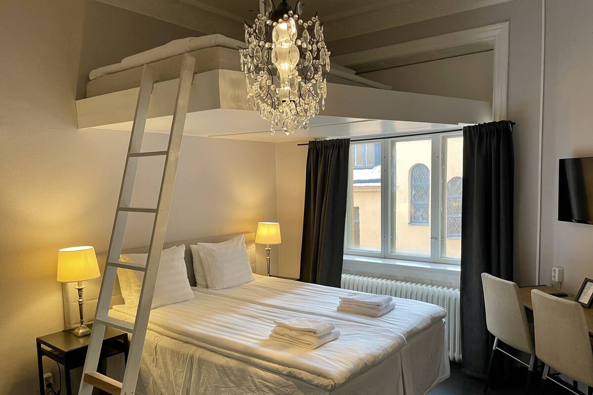 A hotel room with a bunk bed, a chandelier, a desk with a chair, and a view of the outside through a window.
